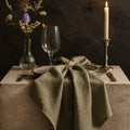 Olivengrøn stofserviet i bomuld og hør, 40x40 cm, med rustik og naturlig tekstur, vist sammenfoldet på spisebordet.
